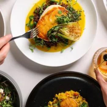 A person uses a fork and holds a cocktail, surrounded by plates of gourmet dishes, including mashed potatoes, steak, vegetables, and colorful entrees on a white table.