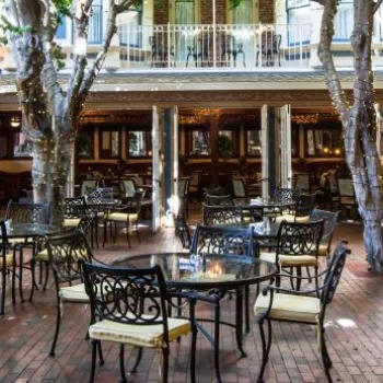 Outdoor patio area with black metal tables and chairs, brick flooring, trees, and surrounding brick walls, set up for dining at a restaurant.