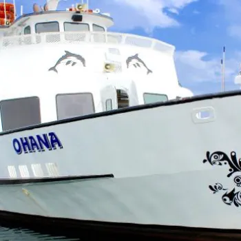 A white boat named OHANA is docked at a marina, with another boat and waterfront buildings visible in the background under a blue sky.