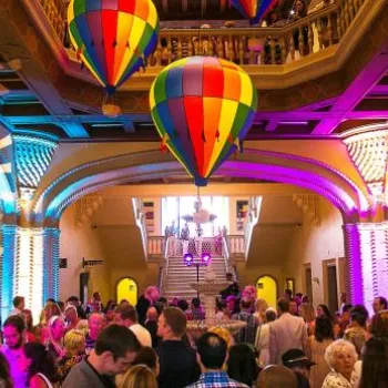 A large crowd gathers in a brightly lit, ornately decorated hall with colorful hot air balloon decorations and light patterns projected on the walls.