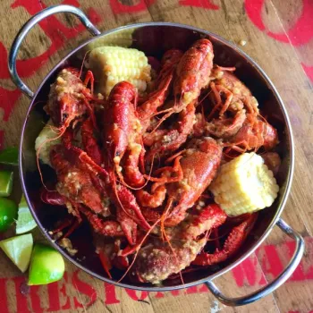 A metal pot filled with cooked crawfish, corn on the cob, and lime wedges, placed on a wooden table.