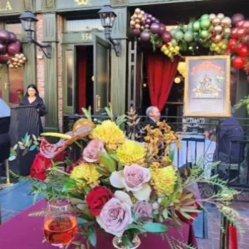 A festive restaurant entrance decorated with colorful balloons and flowers, featuring outdoor seating and a table with a floral arrangement and drinks in the foreground.