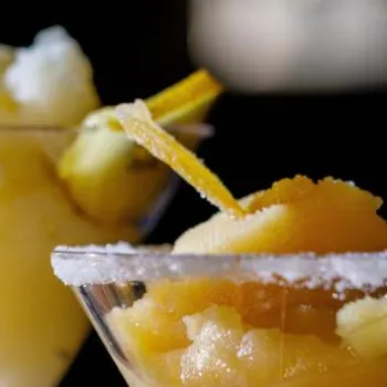 Two martini glasses filled with frozen cocktails, each garnished with a lemon twist and a gold-colored spoon, against a dark background.