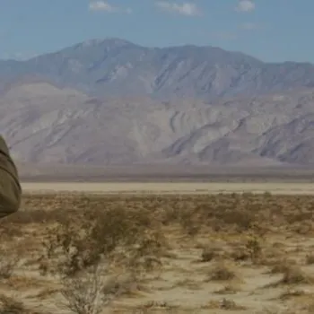 A park ranger wearing a hat looks through binoculars at a dry desert landscape with mountains in the distance.