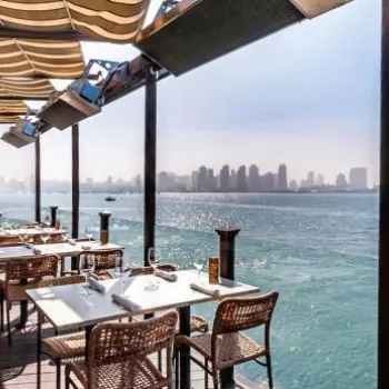 Outdoor restaurant terrace with wicker chairs and set tables overlooks a large body of water, with a city skyline visible in the distance under a clear blue sky.