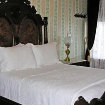 A vintage bedroom with an ornate dark wood bed, white bedding and pillows, lace curtains, striped wallpaper, and a small table with a lamp.