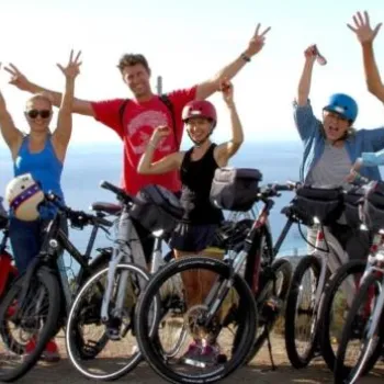 A group of people pose with bicycles outdoors, raising their arms and smiling, with the ocean visible in the background.