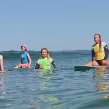 Six people wearing wetsuits sit on surfboards in the ocean, facing the camera, under a clear sky.