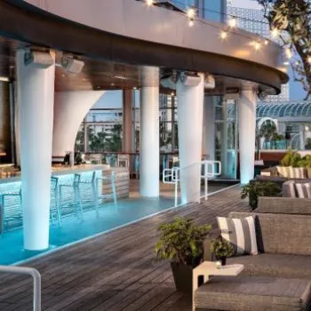 Outdoor rooftop lounge with modern seating, a long bar with stools, string lights overhead, and greenery in the background.