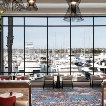 Modern restaurant interior with large windows overlooking a marina with boats; tables and booths are set for dining under contemporary lighting fixtures.