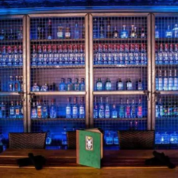 A dimly lit room with shelves of liquor bottles behind metal cages, a wooden table with a green menu, and two black napkins in the foreground.