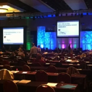 Conference room with rows of tables and chairs, people seated and standing, large projection screens at the front displaying presentation slides.