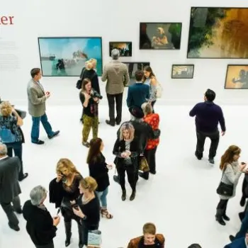 People mingle at an art gallery event with framed photographs on white walls; a live band plays in the corner.