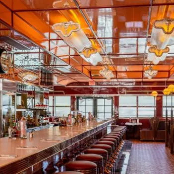 A retro-style diner with red booths, a long counter lined with red stools, art deco lighting, and mirrored walls.