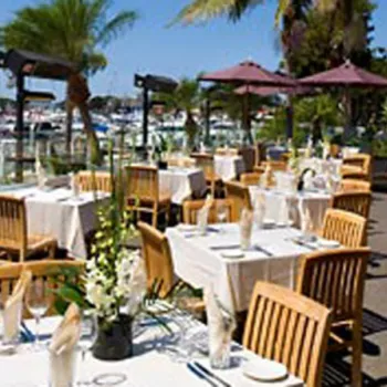 Outdoor restaurant patio with wooden tables and chairs, white tablecloths, umbrellas, and a view of a marina with boats and palm trees in the background.