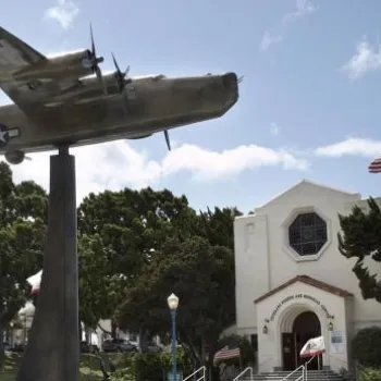 A large aircraft sculpture on a pedestal stands in front of a white building with multiple flags and a clear sky in the background.