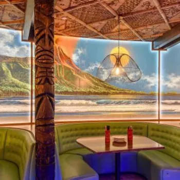 A tropical-themed restaurant interior with green booth seating, wooden tables, hanging lights, and a mural of an island beach and mountains on the wall.