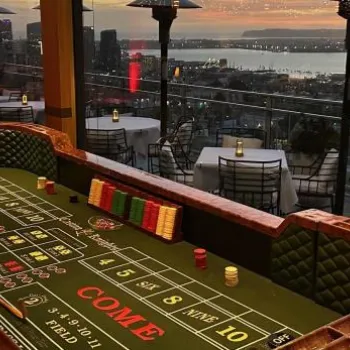 Craps table in a casino room with large windows, outdoor seating, and a cityscape view at sunset over a bay.