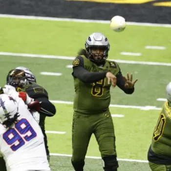 A football quarterback in a green uniform throws a pass while offensive and defensive players block each other on the field.