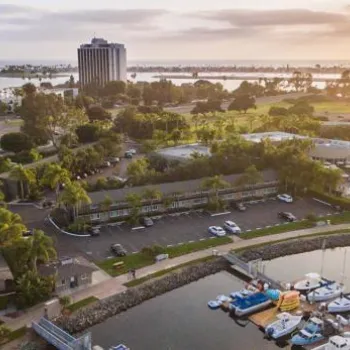 Aerial view of a marina with docked boats, nearby buildings, palm trees, and a distant bay at sunset.