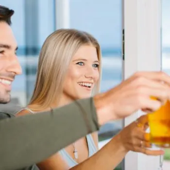Three people sitting indoors clink glasses in a toast, with a beach and ocean visible through the window in the background.