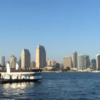 A white ferry approaches a pier with people onboard; San Diego skyline and tall buildings are visible across the bay under a clear blue sky.