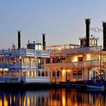 A large, brightly lit riverboat with paddlewheel design is docked by the water at sunset, with small sailboats and palm trees in the background.