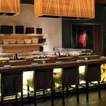 A sushi chef prepares food behind a well-lit sushi bar with several empty chairs; shelves with plates and a bar area are visible in the background.