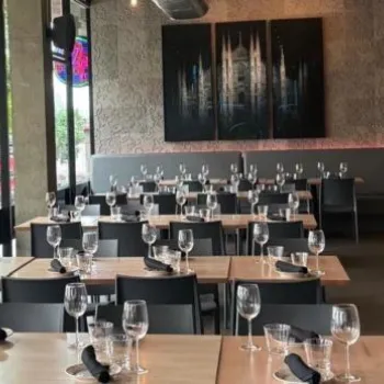 Modern restaurant dining area with neatly arranged tables, black chairs, glassware, and napkins, large windows, wall art, and potted plants.