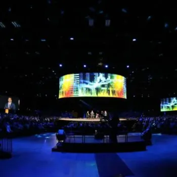 A large audience sits in a dark auditorium facing a brightly lit stage with a wide, curved screen displaying colorful graphics. Multiple smaller screens flank the main stage.