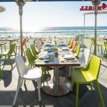 Seaside restaurant terrace with colorful chairs and set tables overlooking a busy beach and ocean under a clear sky.
