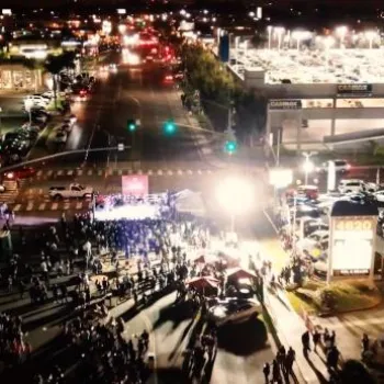 A large crowd gathers in a shopping center parking lot at night, with bright lights illuminating the area and nearby stores and cars visible.