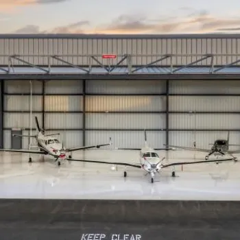 Two small airplanes are parked inside a clean, open aircraft hangar at sunset, with a "KEEP CLEAR" sign visible on the ground outside.