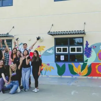 A group of people pose and celebrate in front of a building with a colorful mural that reads "GROW YOUR DREAMS" and features flowers and a hummingbird.