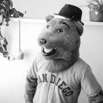 A person in a bear mascot costume wearing a "San Diego" shirt and a hat stands indoors near plants.