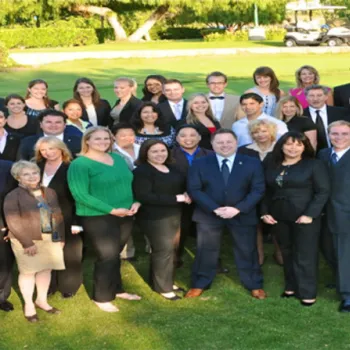 A group of professionally dressed adults poses outdoors on a lawn, standing in rows and smiling at the camera. Trees and a golf cart appear in the background.