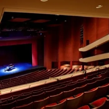 An empty theater with rows of red seats faces a stage with a piano under spotlights; balconies and lighting fixtures are visible.