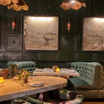 Elegant restaurant interior with plush green booths, marble tables set for dining, framed art on dark paneled walls, soft lighting, and decorative pendant lights overhead.