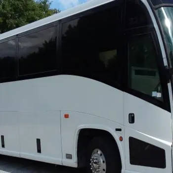 A large white coach bus is parked on a paved surface near trees with partly cloudy skies in the background.