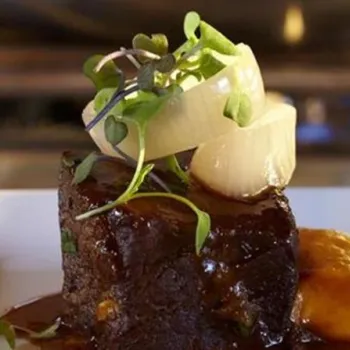 A square portion of braised meat with dark sauce, topped with onion rings and microgreens, served with a dollop of orange puree on a white plate.