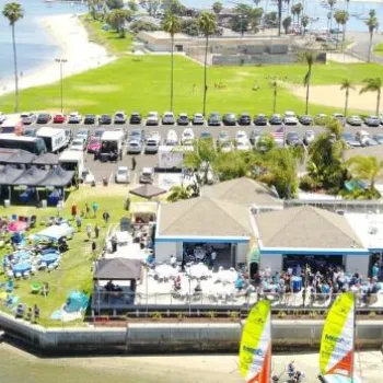 Aerial view of a waterfront event with crowds, tents, parked cars, colorful sailboats, and palm trees on a grassy area near the beach.