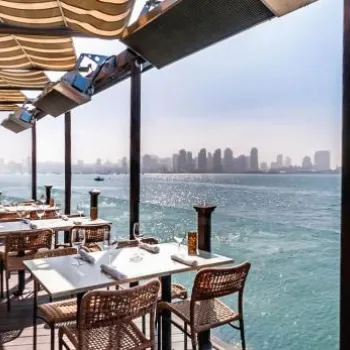 Outdoor restaurant terrace with tables and chairs overlooking a calm blue body of water and a distant city skyline under a clear sky.