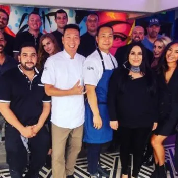 A group of restaurant staff poses together indoors, standing in front of a colorful mural, smiling at the camera.