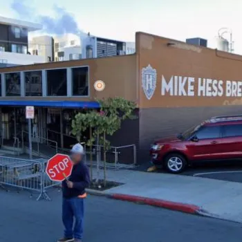 A person holding a stop sign stands in front of Mike Hess Brewing; several cars are parked alongside the building on a sunny day.