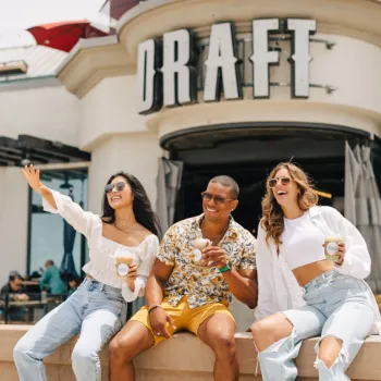 Three people sit on a ledge outside a building with a "DRAFT" sign, smiling and holding drinks, with two women wearing sunglasses and casual outfits and the man in shorts and a shirt.