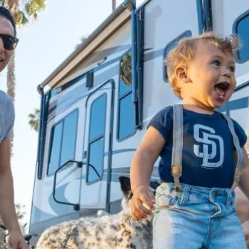 A man, woman, and young child are outside near an RV on a sunny day. The child is smiling and wearing a blue shirt while the adults are sitting and standing nearby.