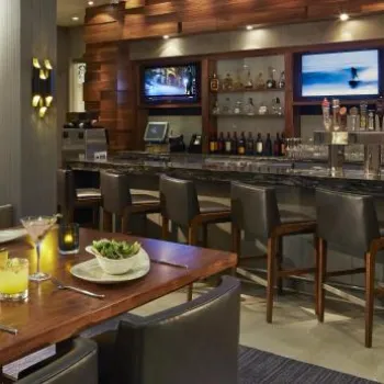 Modern bar and restaurant with a stocked bar, high chairs, TV screens, and a dining table set with drinks, salads, and appetizers. Dim, ambient lighting throughout the space.