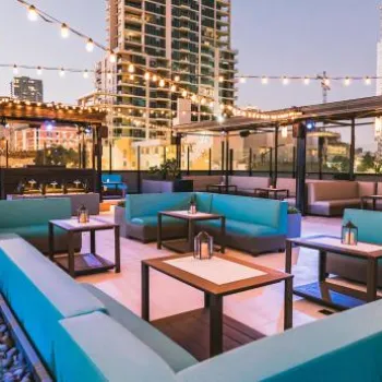Rooftop lounge with turquoise sectional seating, string lights overhead, and city buildings in the background at dusk.