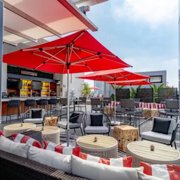 Outdoor rooftop lounge with red umbrellas, cushioned seating, round tables, and a bar area in the background under a partly cloudy sky.