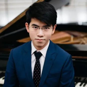 A young man in a blue suit and glasses sits in front of a grand piano, looking directly at the camera.